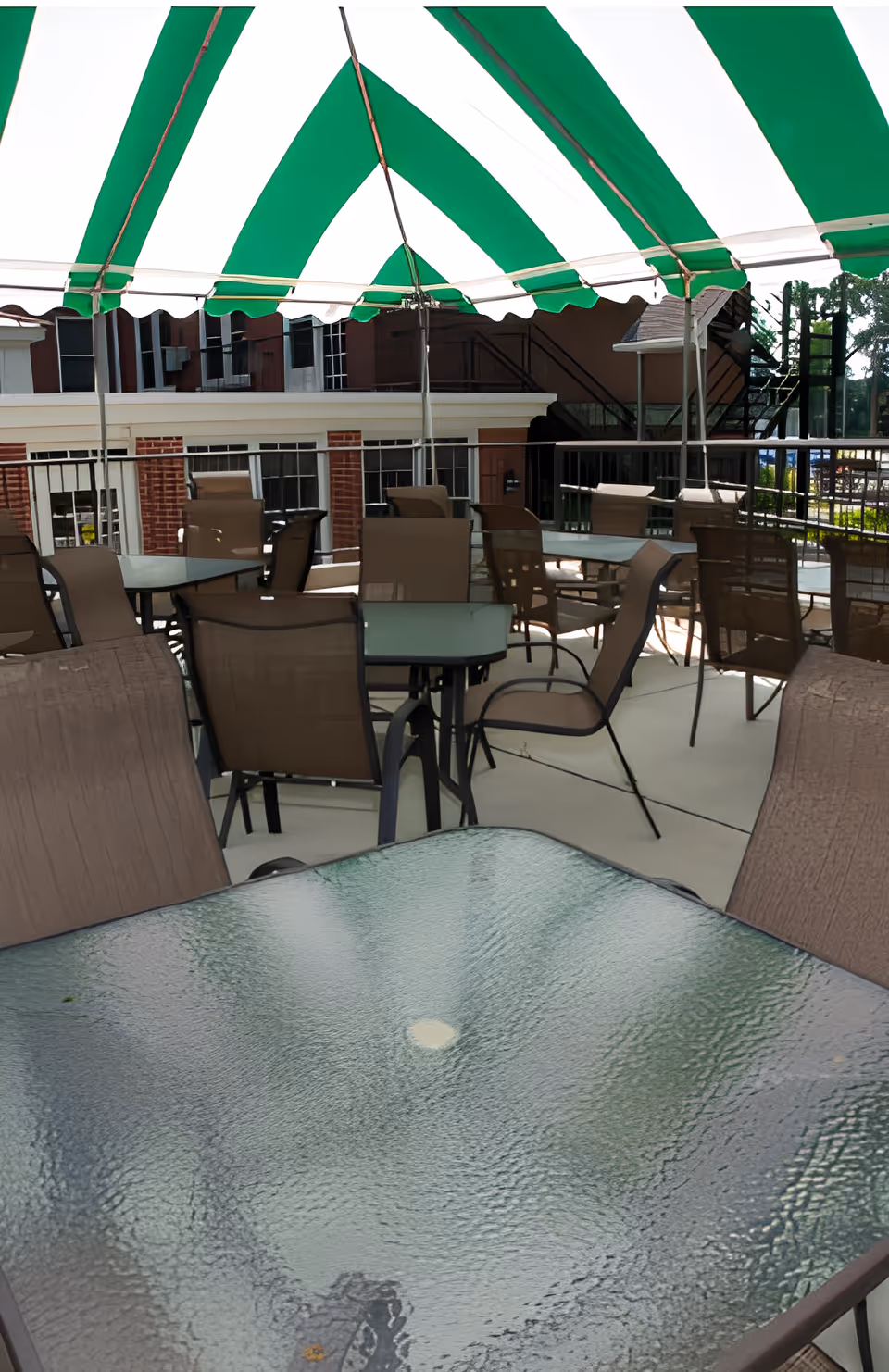 Outdoor seating area with multiple glass-top tables and brown mesh chairs under a green and white striped canopy. The area is surrounded by a metal railing and there are buildings and stairs visible in the background.
