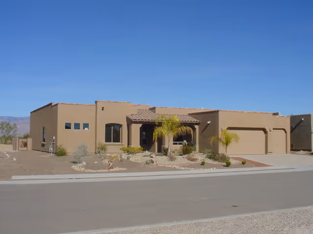 Single-story tan stucco building with a tiled roof and a three-car garage, surrounded by desert landscaping with small palm trees and rocks under a clear blue sky.