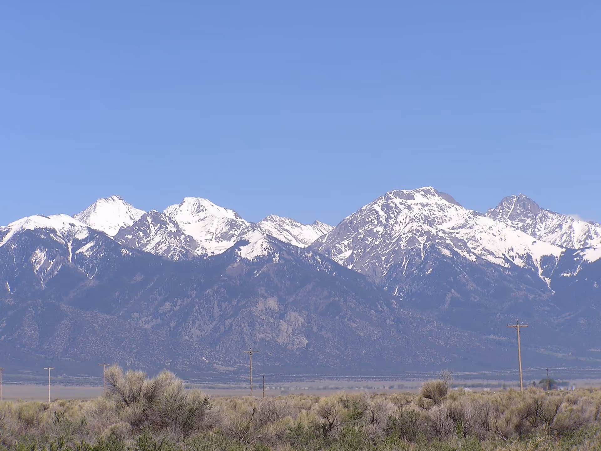 Snow-capped mountains overlook a dry desert plain with scrub and blue sky.