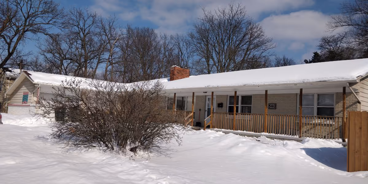 Snow-covered single-story Divine Manor building with a wooden porch and bare trees in background