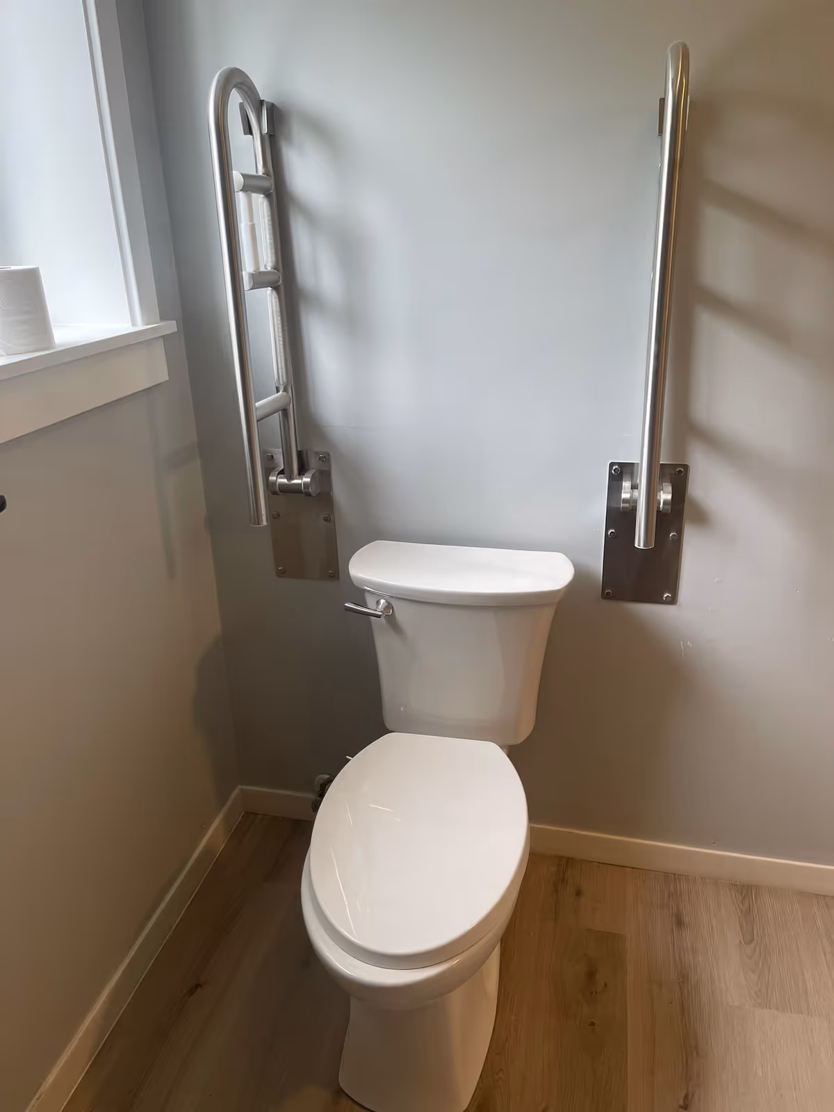 Accessible bathroom with toilet, wall-mounted grab bars, and wood flooring.