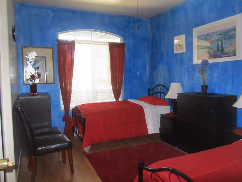 A bedroom with bright blue textured walls and two single beds with red bedding. There is a window with red curtains letting in natural light. The room has wooden flooring, a red rug, two black dressers with lamps and decorative items, and two black chairs against one wall.