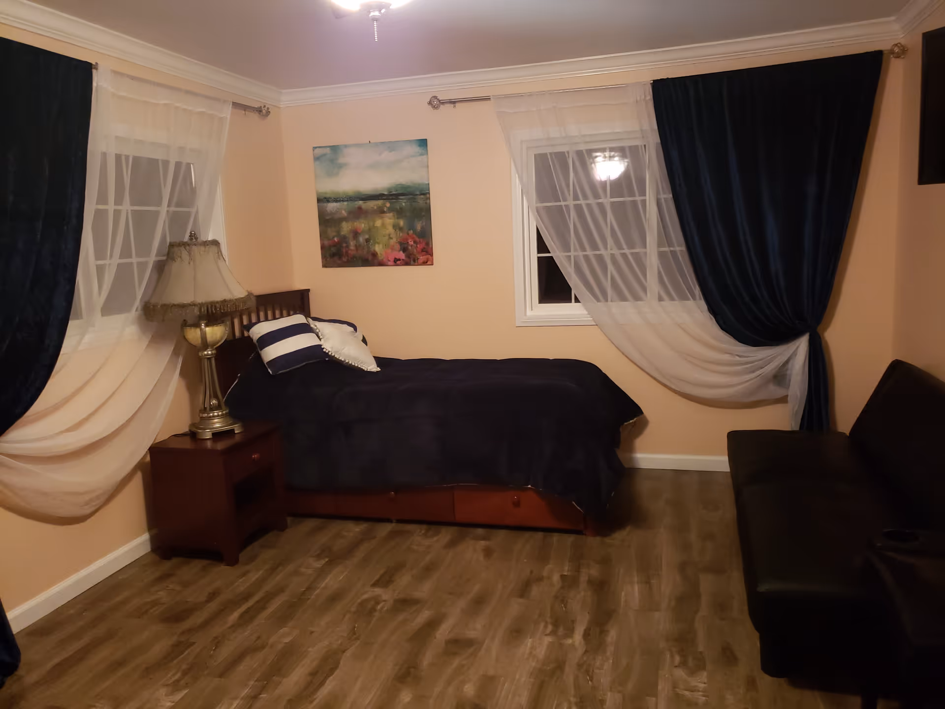 A bedroom featuring a bed, nightstand, lamp, windows with sheer and navy curtains, and a sofa.