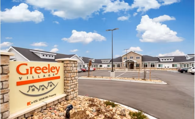 Exterior view of Greeley Village senior living facility with a large parking lot, stone pillars, and a sign displaying the facility name. The building has a modern design with multiple windows and a clear blue sky with some clouds above.