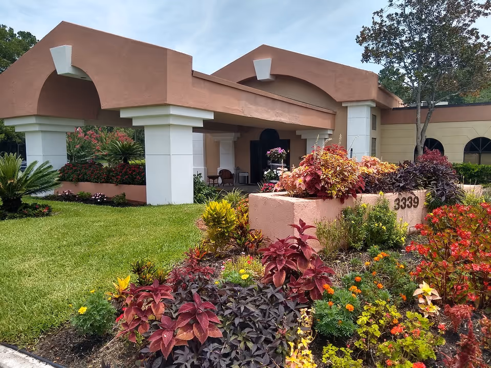 Exterior view of a building entrance with a covered driveway supported by white columns. The building has a tan and brown color scheme. In front, there is a well-maintained garden with various colorful plants and flowers, and a planter with the number 3339 displayed on it.