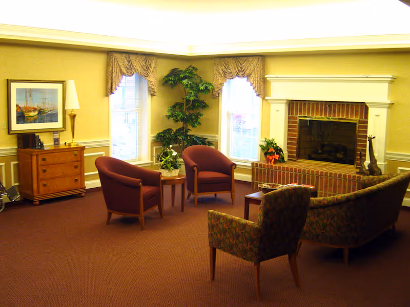 A cozy living room featuring two burgundy armchairs and a patterned sofa around a coffee table, with a brick fireplace and large windows dressed with curtains.