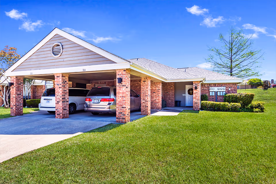Exterior view of a single-story brick building with a covered carport housing two vehicles. The building has a well-maintained lawn, a tree, and a clear blue sky in the background. The carport has numbered spaces 703 and 704.