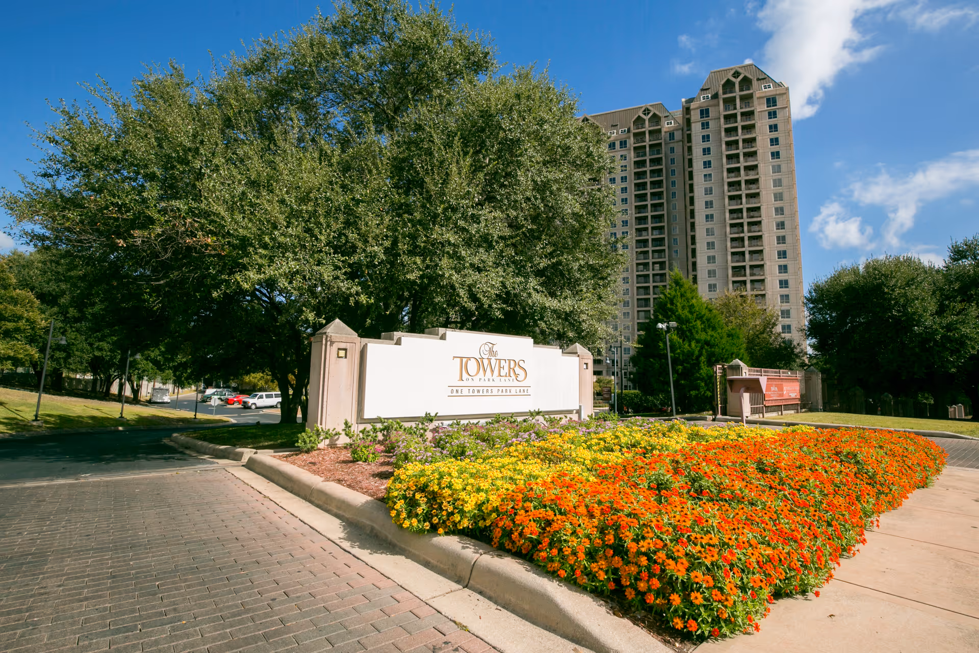 The Towers on Park Lane sign with a tall high-rise building and colorful flower beds.