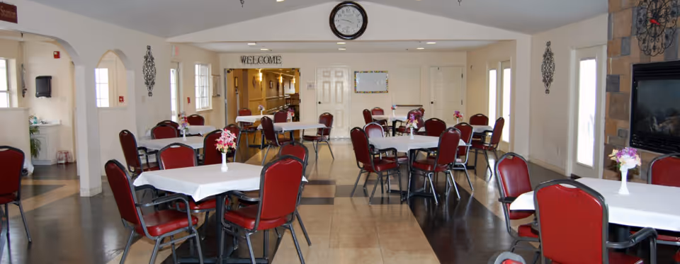 A spacious dining room with multiple tables covered with white tablecloths and decorated with small flower vases. The room has red cushioned chairs around the tables, a clock on the wall above a 'WELCOME' sign, and a stone fireplace on the right side. There are windows and doors letting in natural light.
