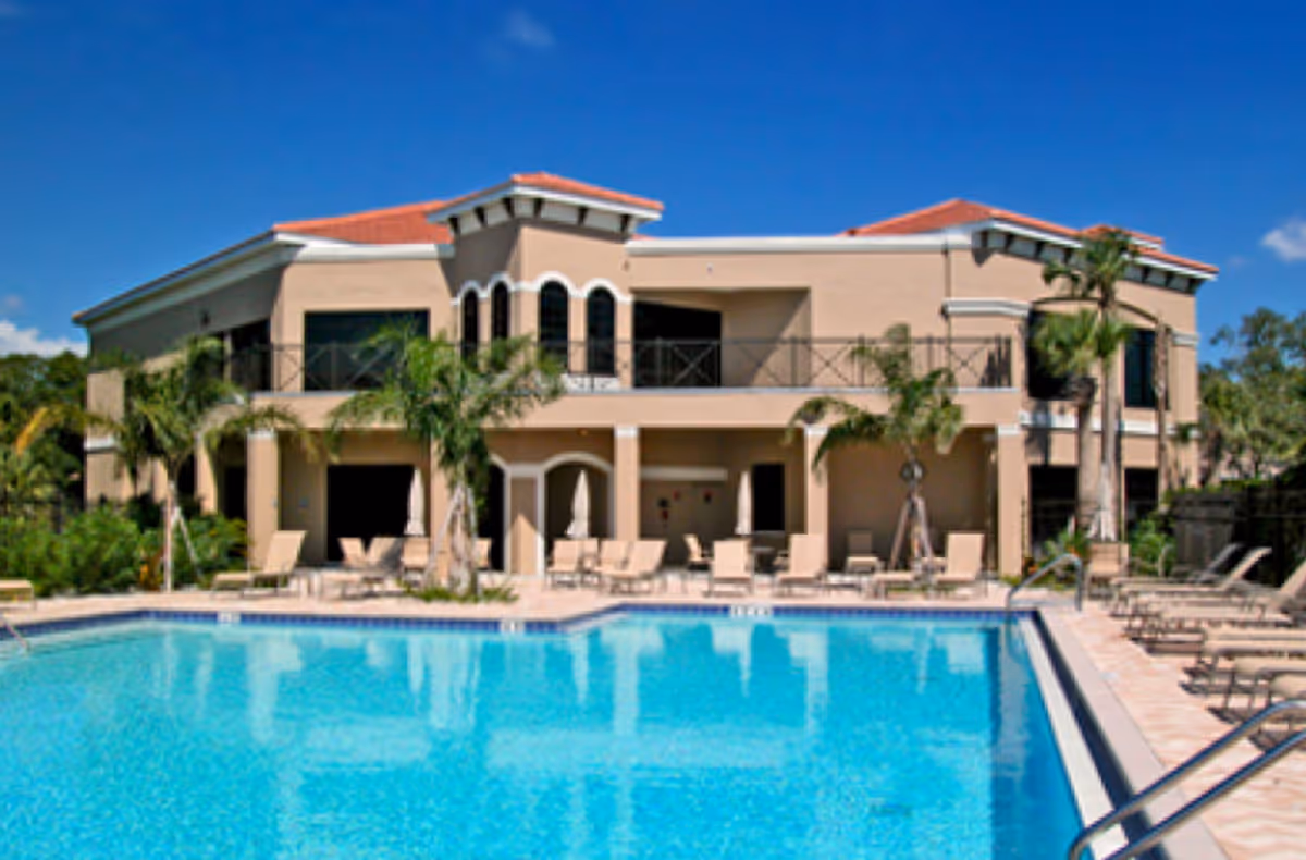 Two-story beige senior living building with a red-tiled roof, palm trees and an outdoor swimming pool with lounge chairs in front.