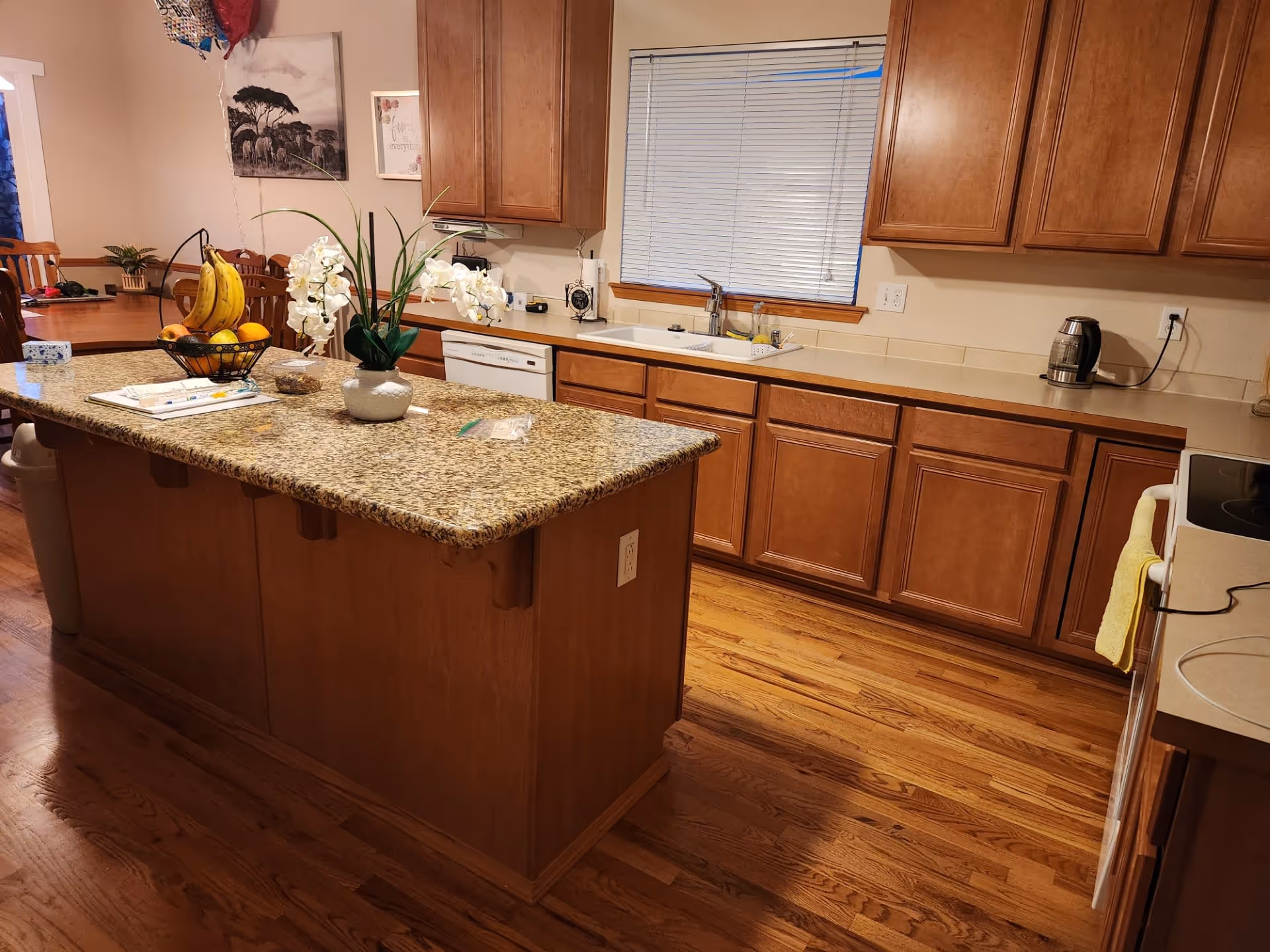 Kitchen featuring a central granite island with fruit and a vase, wooden cabinets and drawers, a sink beneath a window, and hardwood floors.