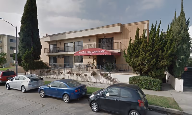 Two-story beige building with a red awning reading 'Beverly Hills Loving Care' and parked cars along the street.