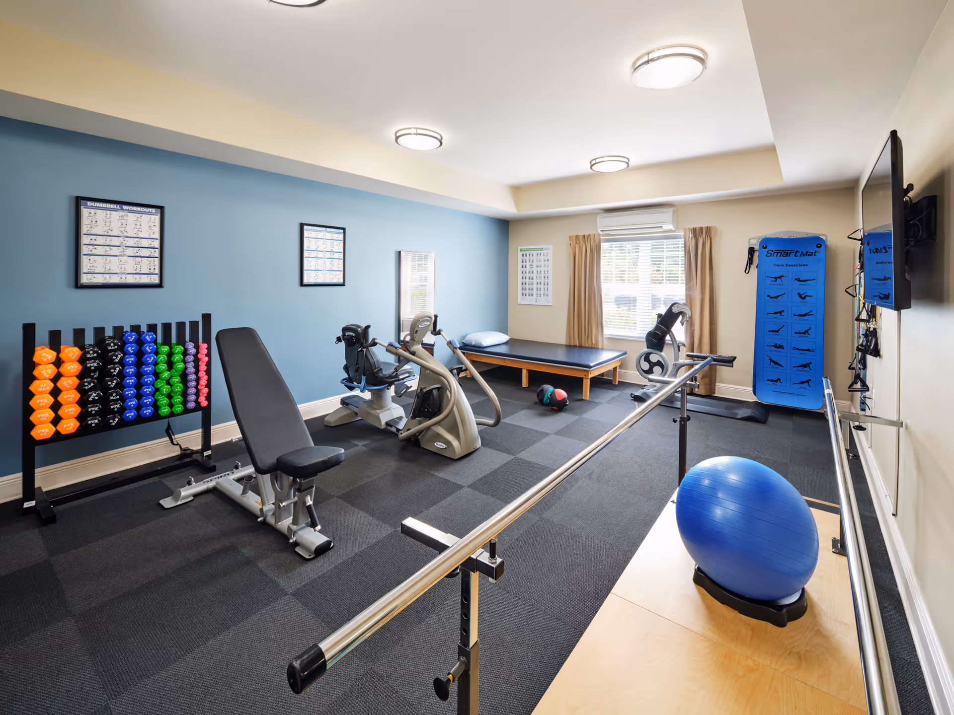 Indoor fitness room with dumbbells, cardio machines, a balance bar, blue wall, and windows.