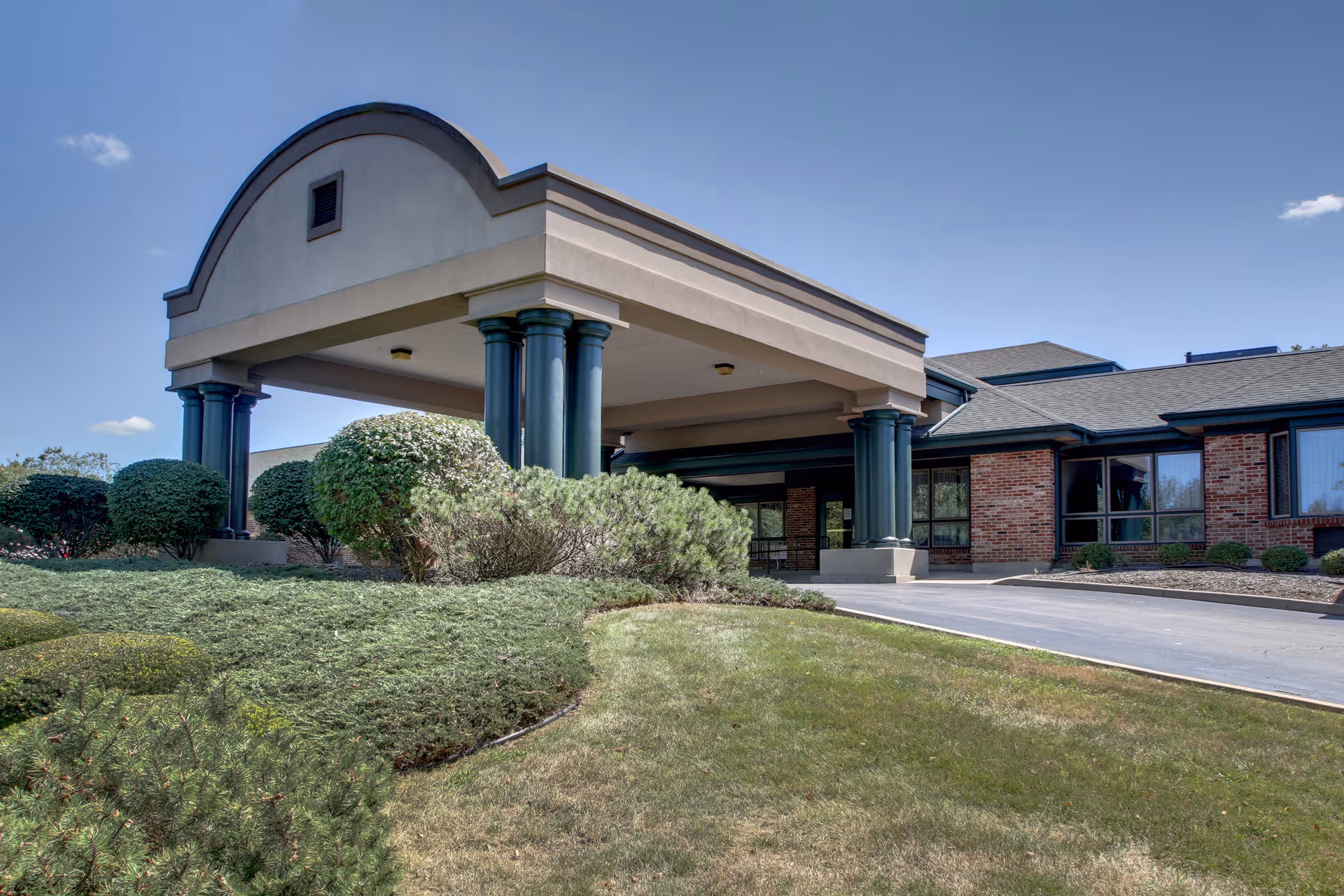 Exterior view of Regency Care facility showing the entrance with a covered drop-off area supported by dark green columns, surrounded by well-maintained bushes and grass under a clear blue sky.