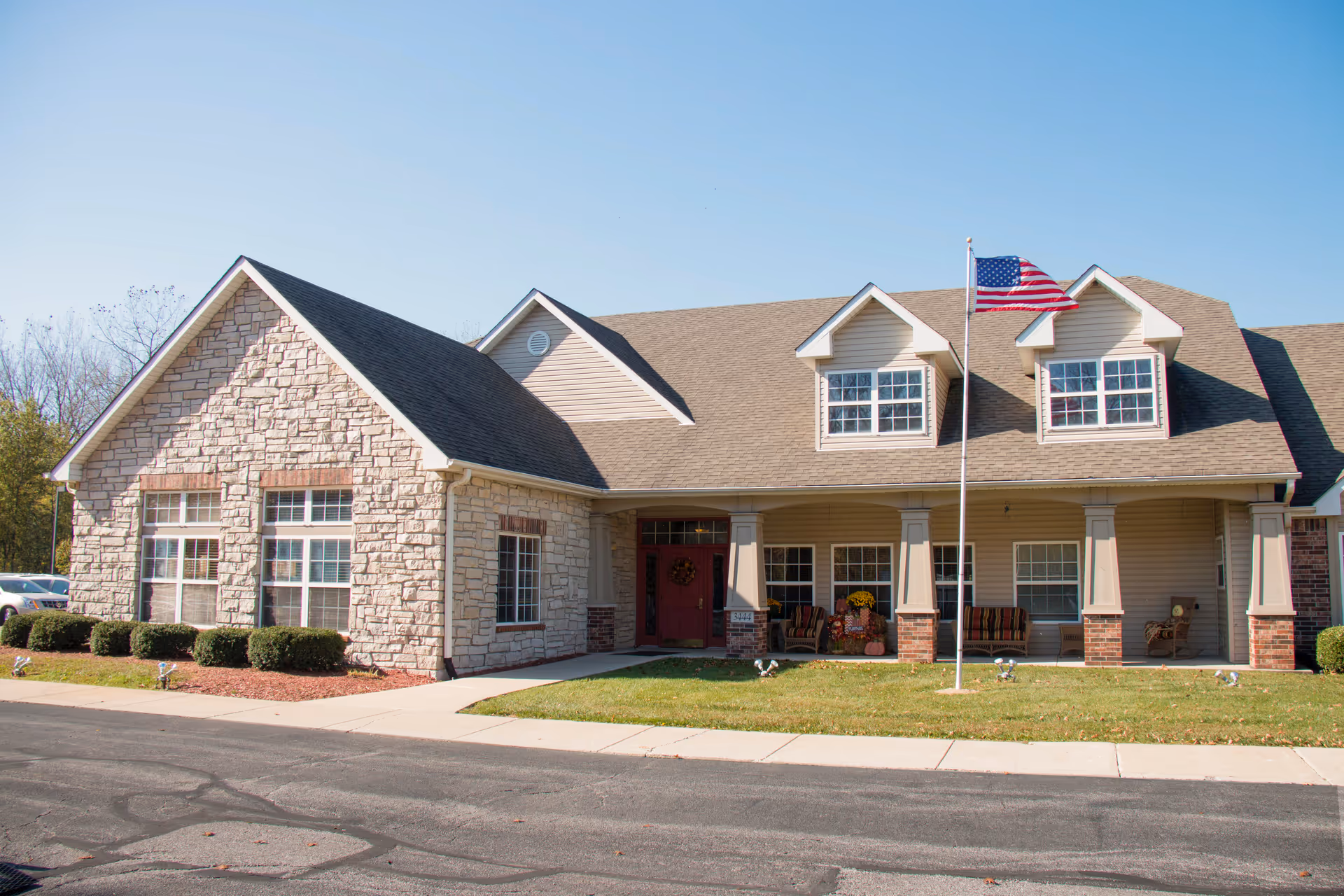 A senior living facility exterior with stone facade, porch seating, and an American flag.