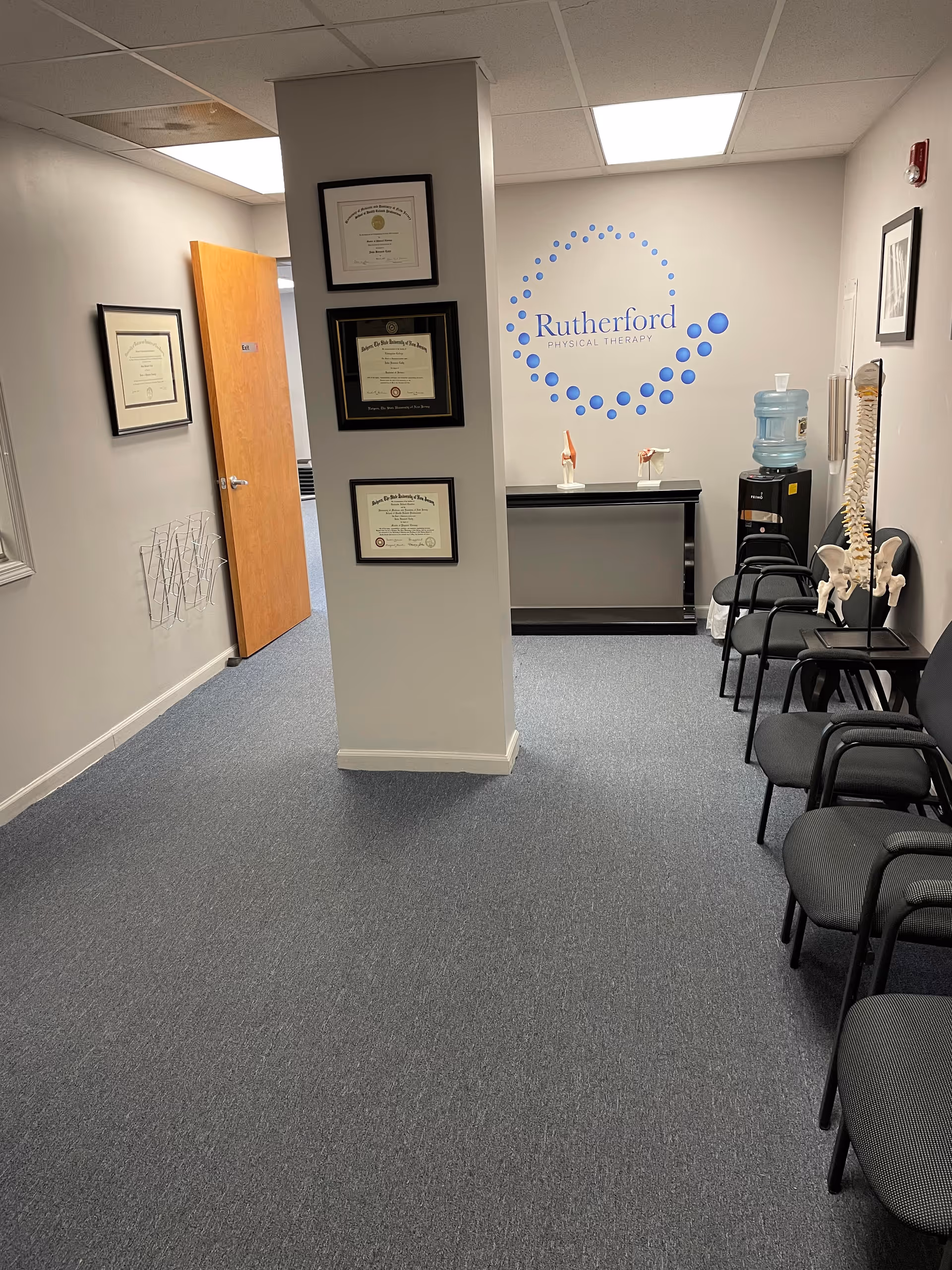 Interior hallway of Rutherford Physical Therapy with wall certificates, chairs, and a water cooler.