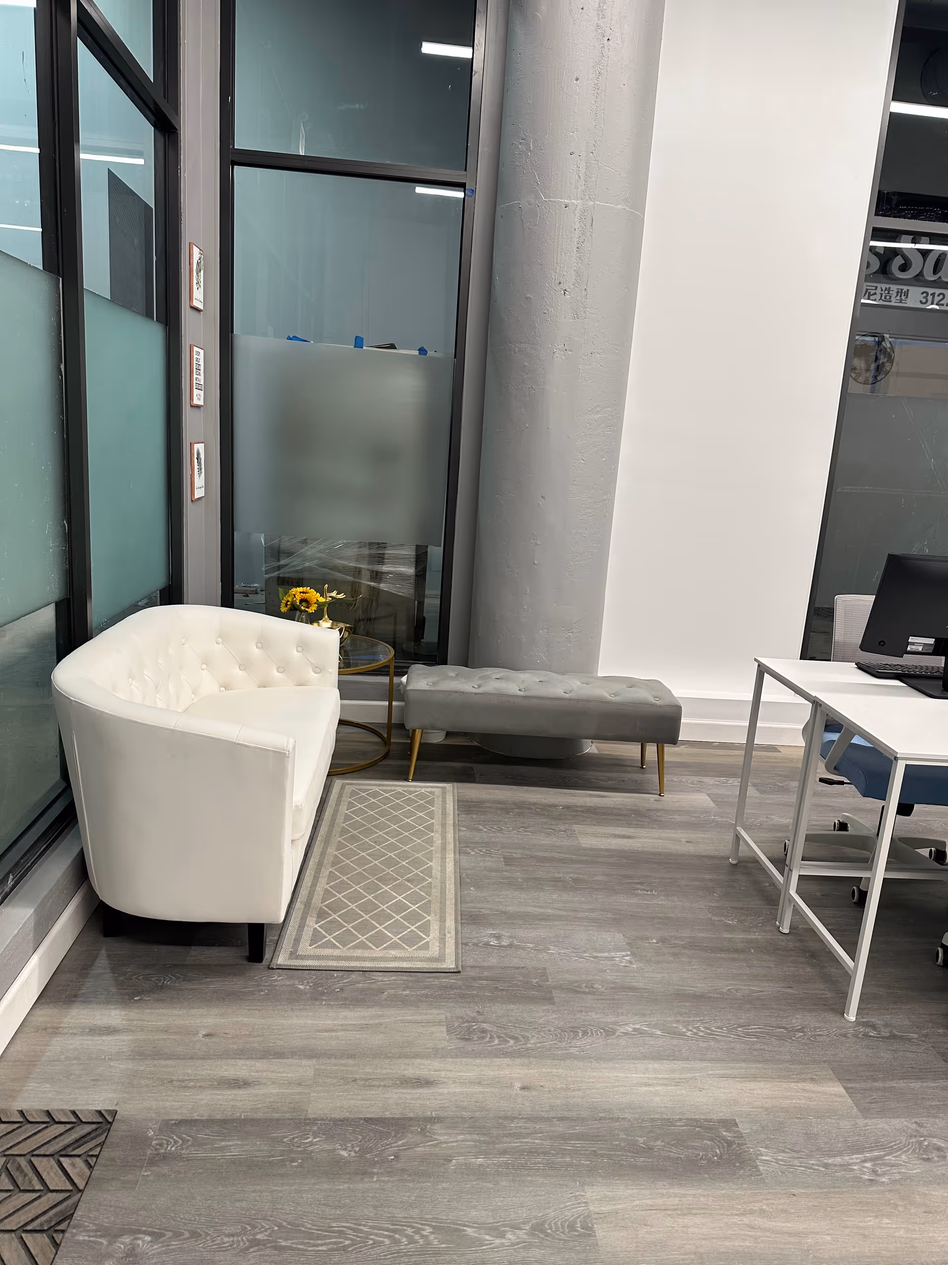 Modern lobby seating area with white tufted chair, gray bench, and desk by glass windows.