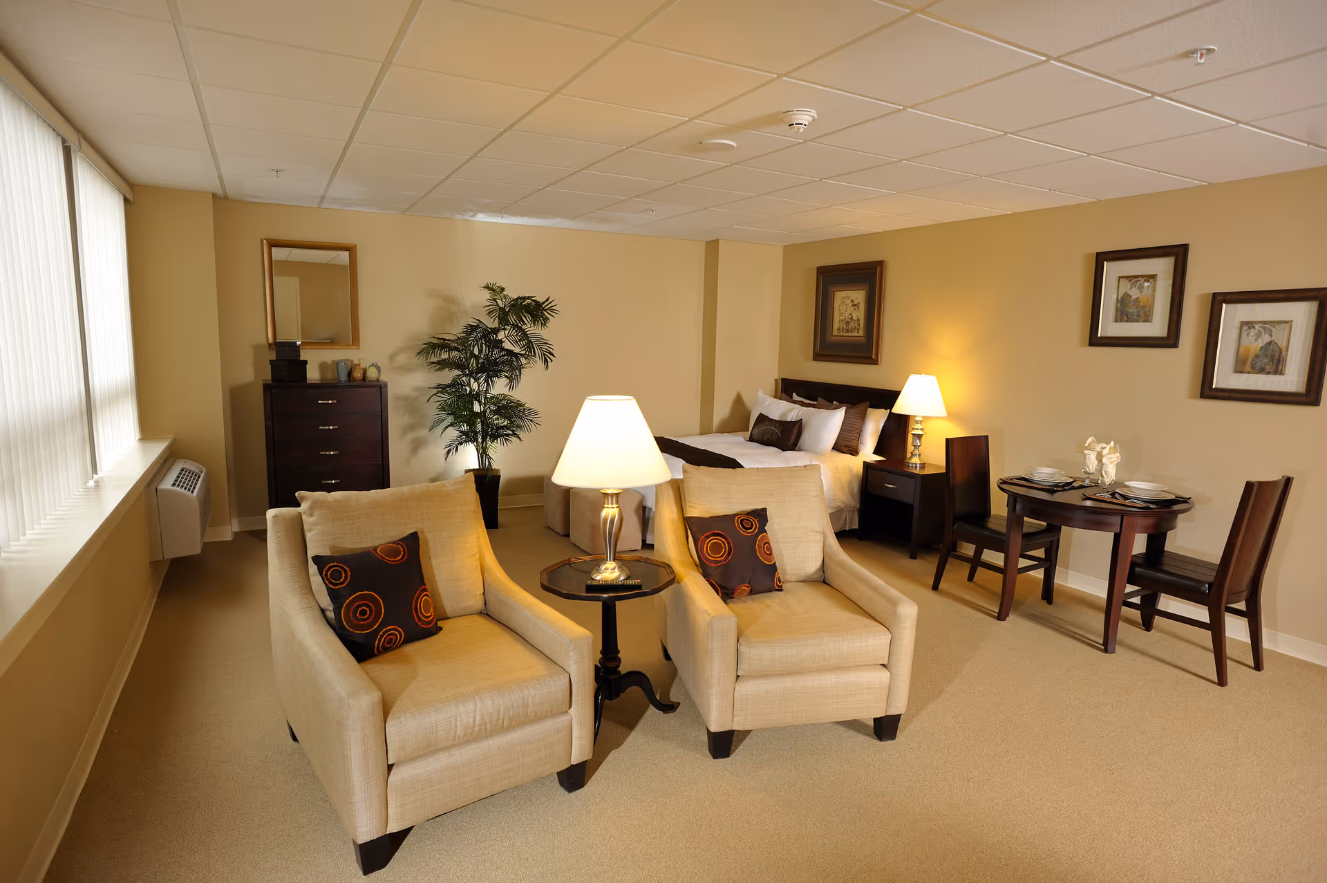 Cozy living area with two beige armchairs, a lamp, table, and adjoining bedroom in a suite.