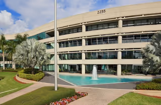 Modern three-story facility exterior with a central pool, palm trees, and landscaped walkway.