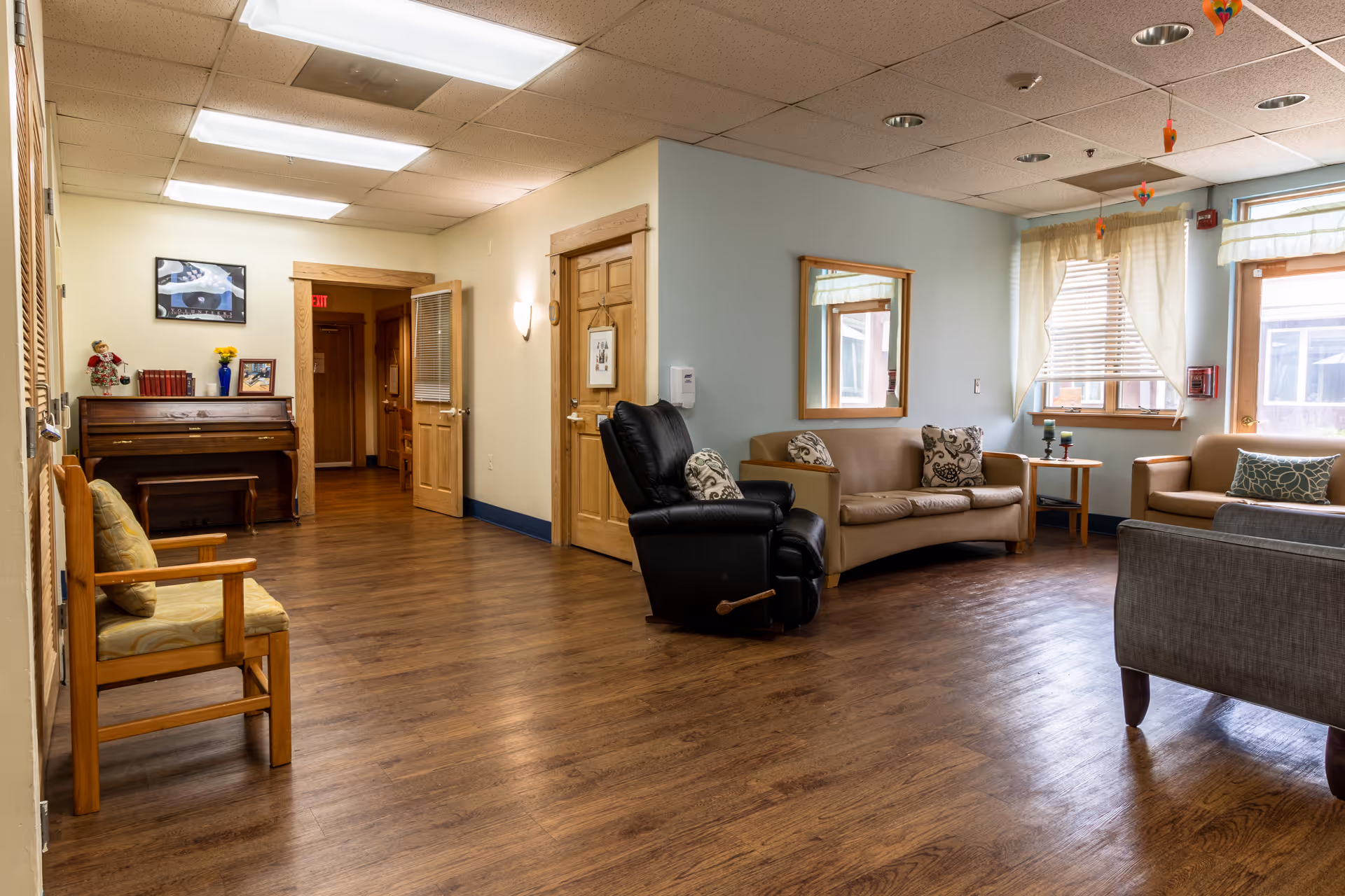 Spacious memory care living room with sofas, a recliner, piano, and large front-facing windows.