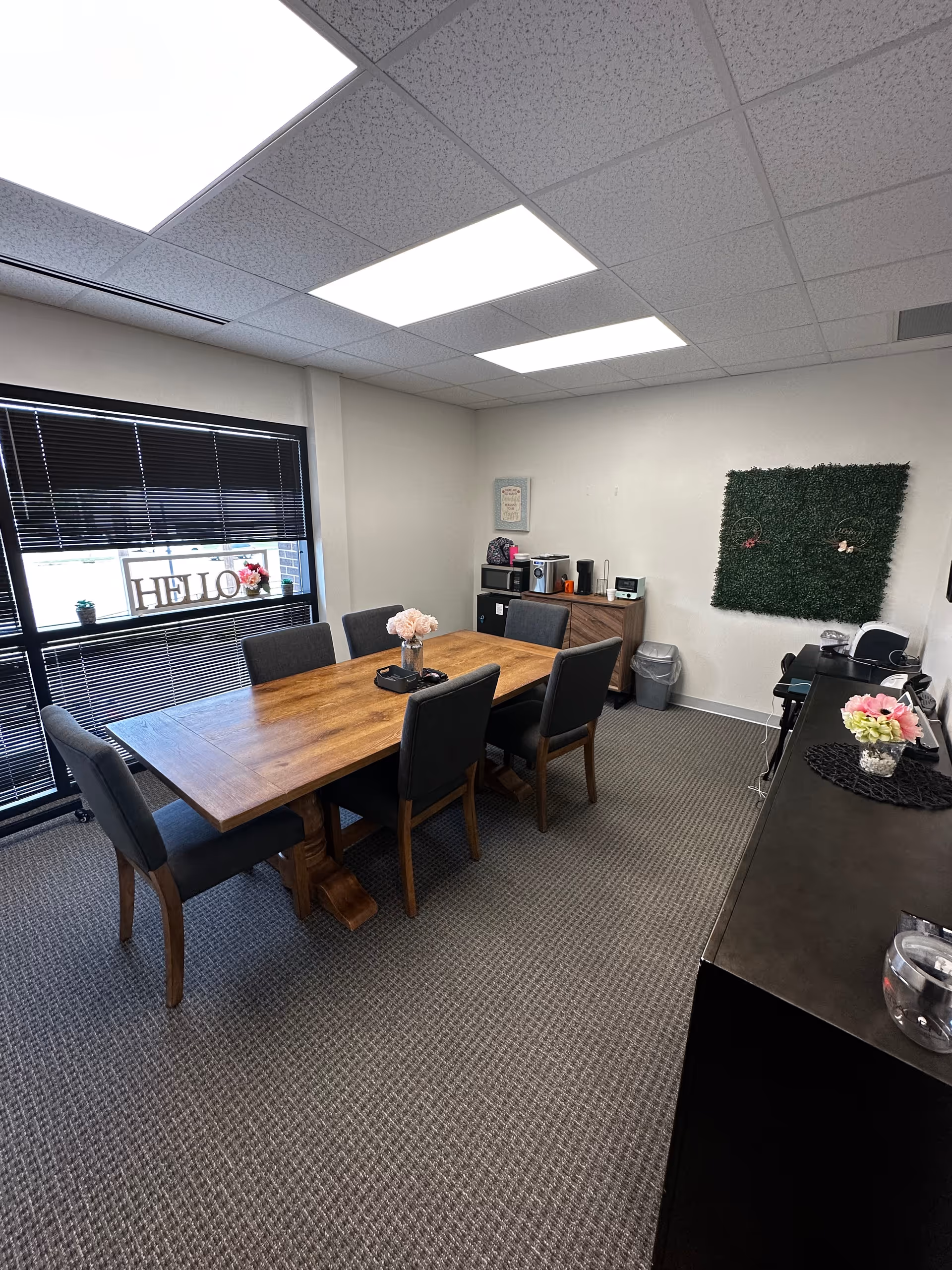 A conference-style dining room with a wooden table, chairs, coffee station, and window view.
