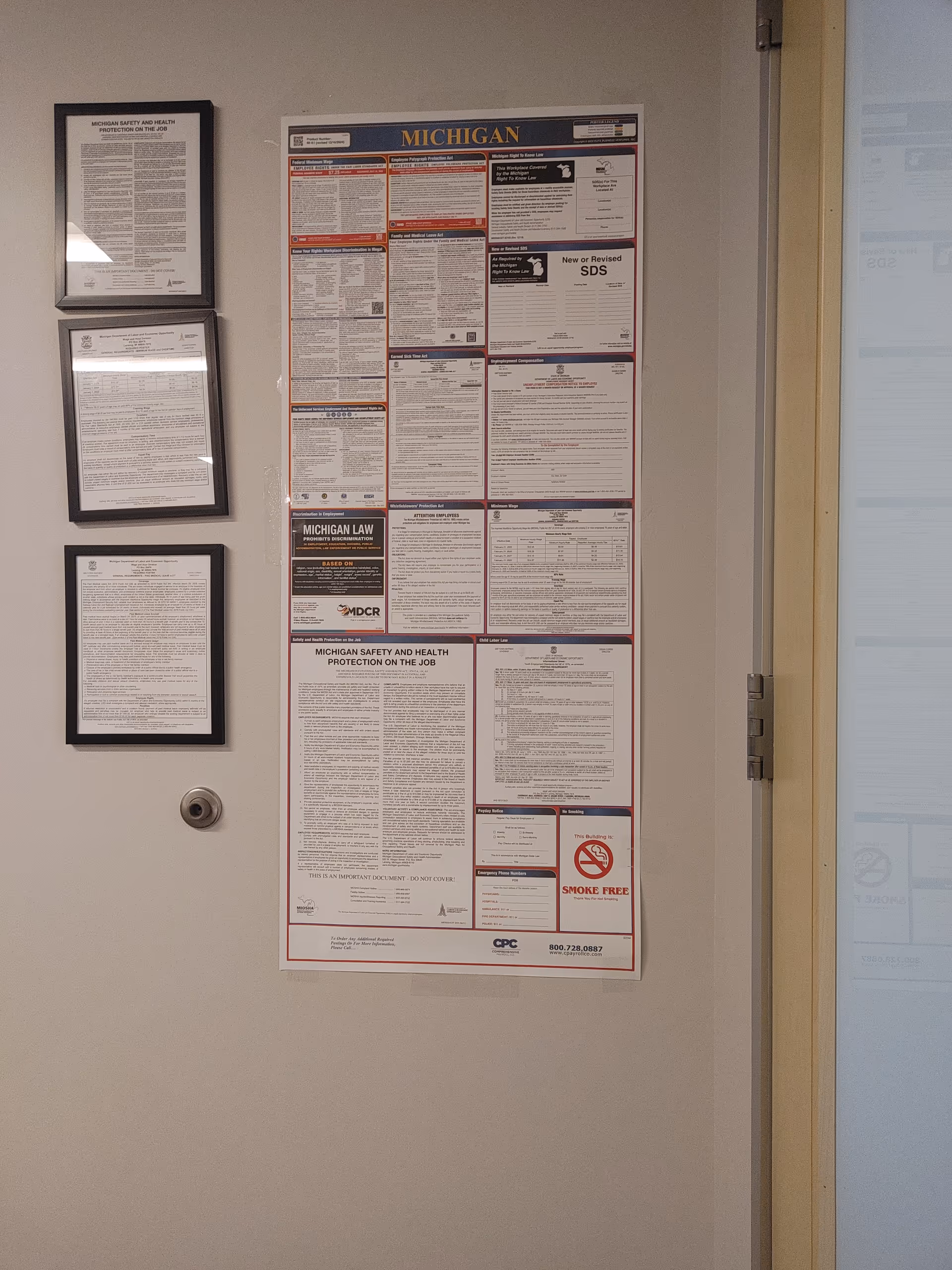 Interior wall with multiple posted notices and framed documents near a door.