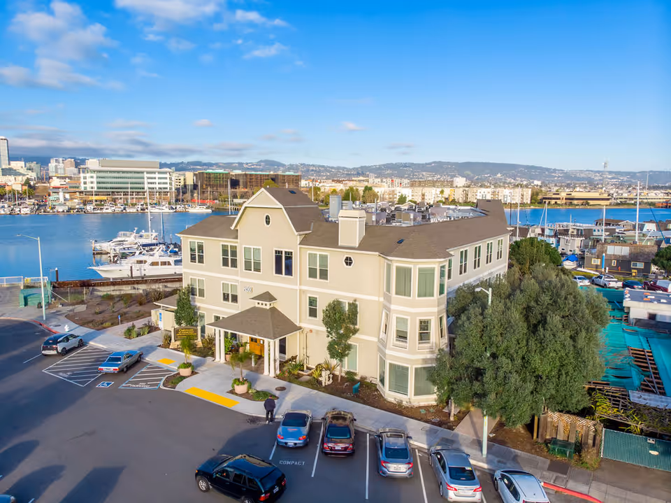 Beige multi-story senior living building along a marina, with parked cars in front.