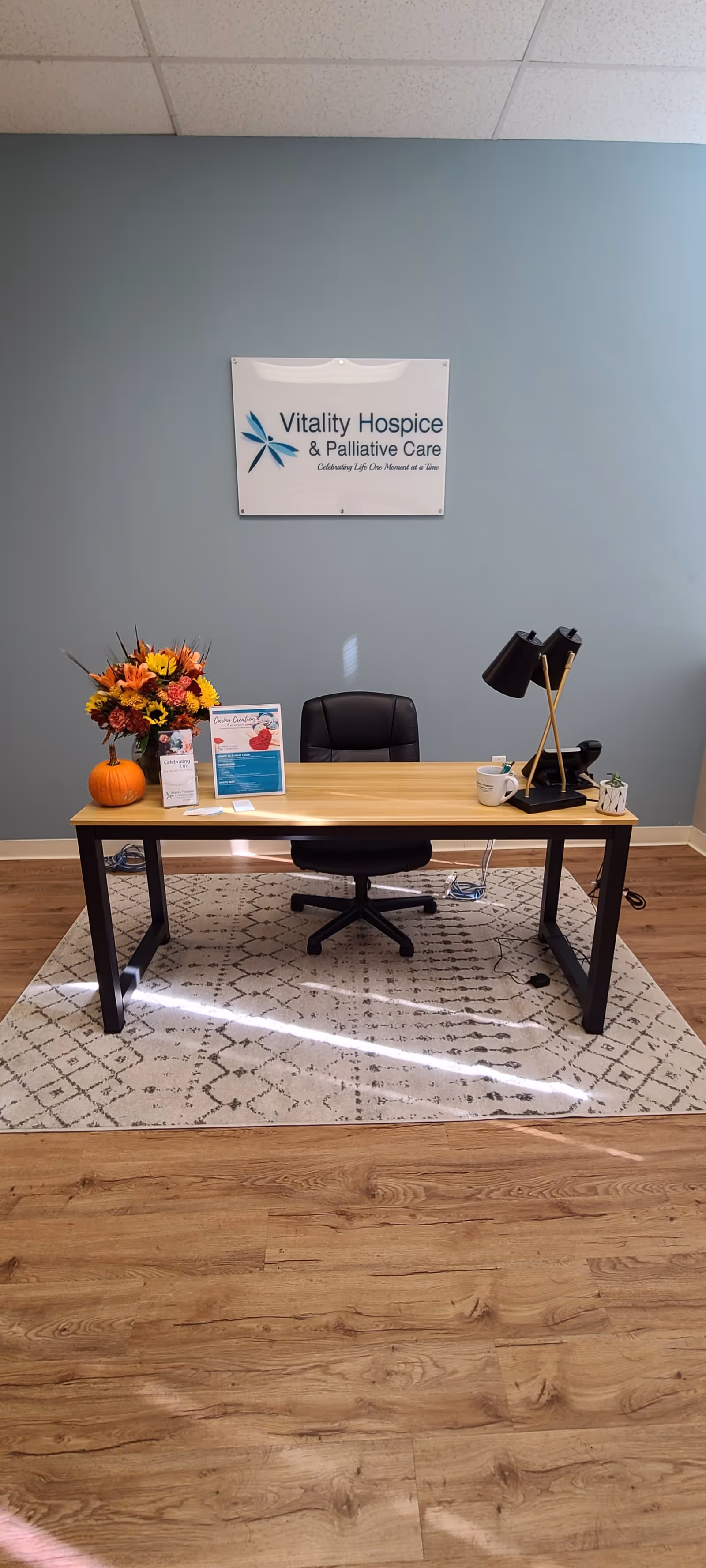 A reception desk with chair, lamp, flowers, brochures, and Vitality Hospice sign on a blue wall.