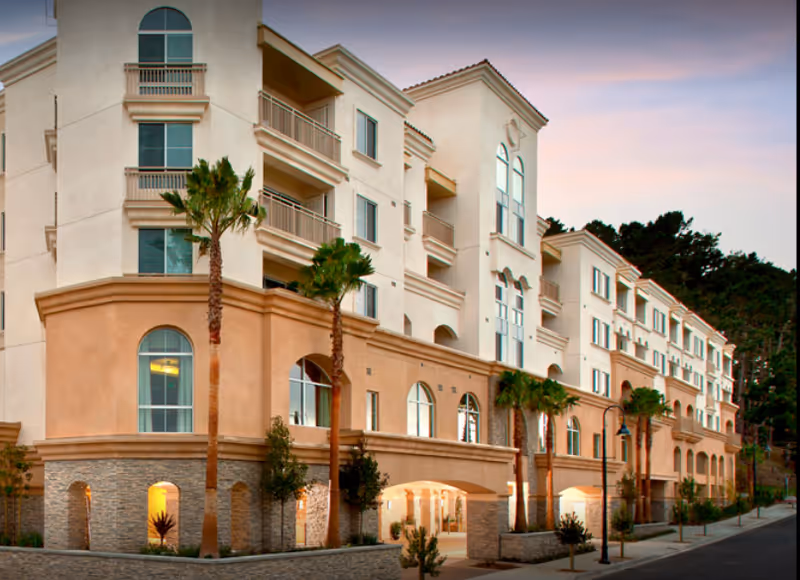 A multi-story senior living facility exterior with beige walls, arches, and palm trees along the street.
