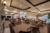Spacious senior living common dining and lounge area with multiple tables and chairs, a stone accent wall, coffered ceiling, and warm lighting.