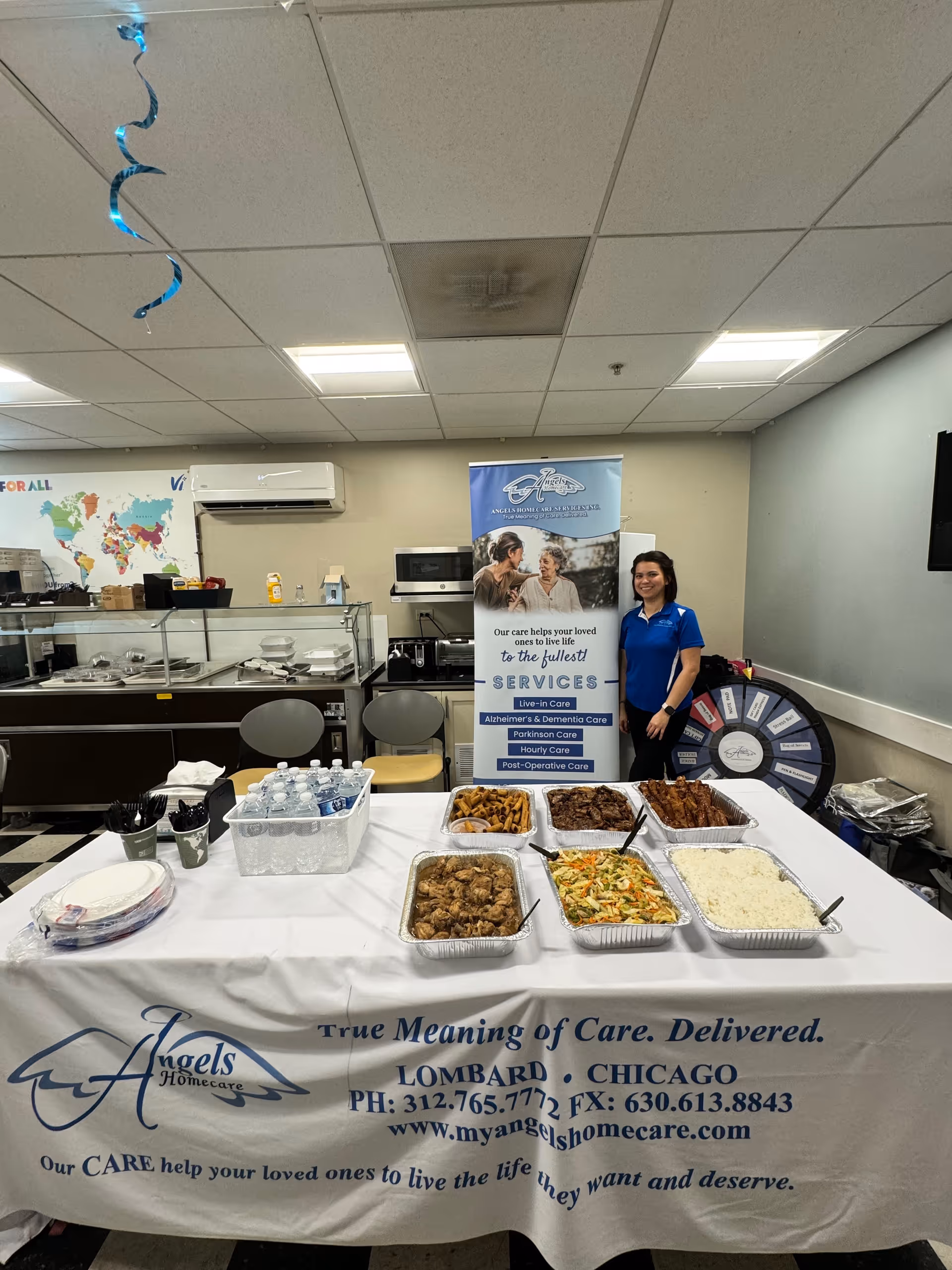 Indoor dining setup with Angels Homecare banner, food trays, and a staff member nearby.