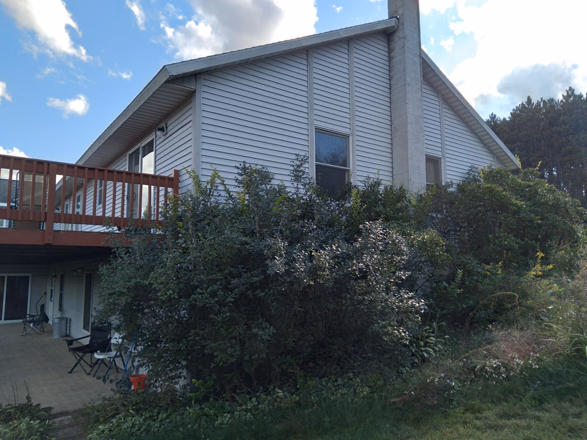 Exterior view of a two-story building with light gray siding and a chimney. The building has a wooden balcony on the upper floor and a covered patio area below with chairs and a table. There are dense bushes and greenery in front of the building, and a partly cloudy sky is visible above.