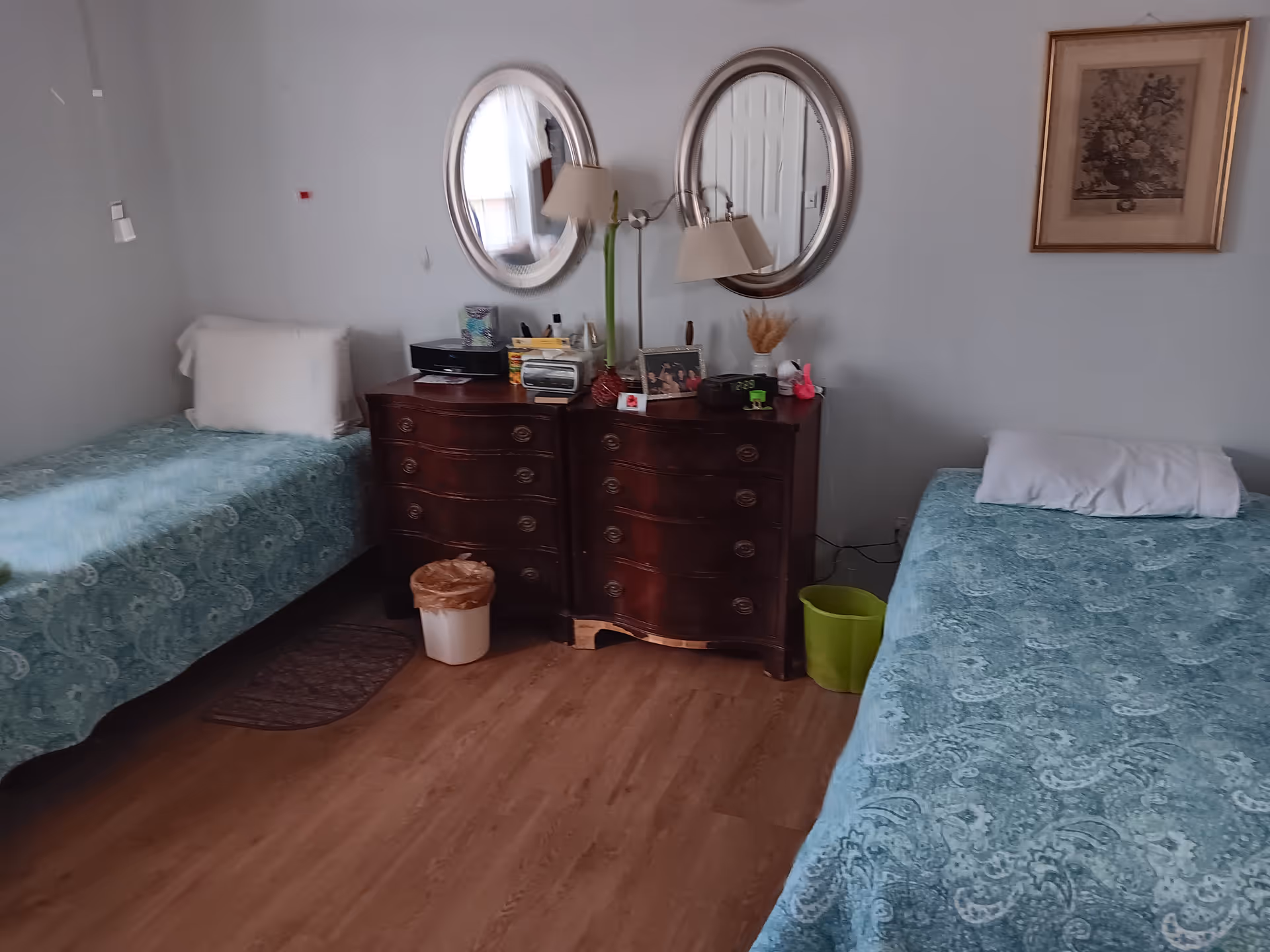Two twin beds with teal patterned bedspreads flank a dark wood dresser beneath two round mirrors.