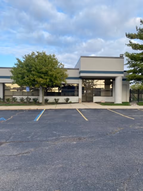 Front view of a modern clinic with glass doors, trees, and a parking lot.