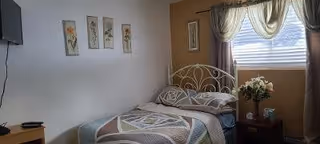 A small bedroom with a single bed featuring a metal headboard, patterned bedding, and two pillows. On the wall above the bed are four framed floral artworks. A window with sheer curtains allows natural light into the room. Next to the bed is a wooden nightstand with a vase of flowers and a small lamp. A flat-screen TV is mounted on the wall opposite the bed.