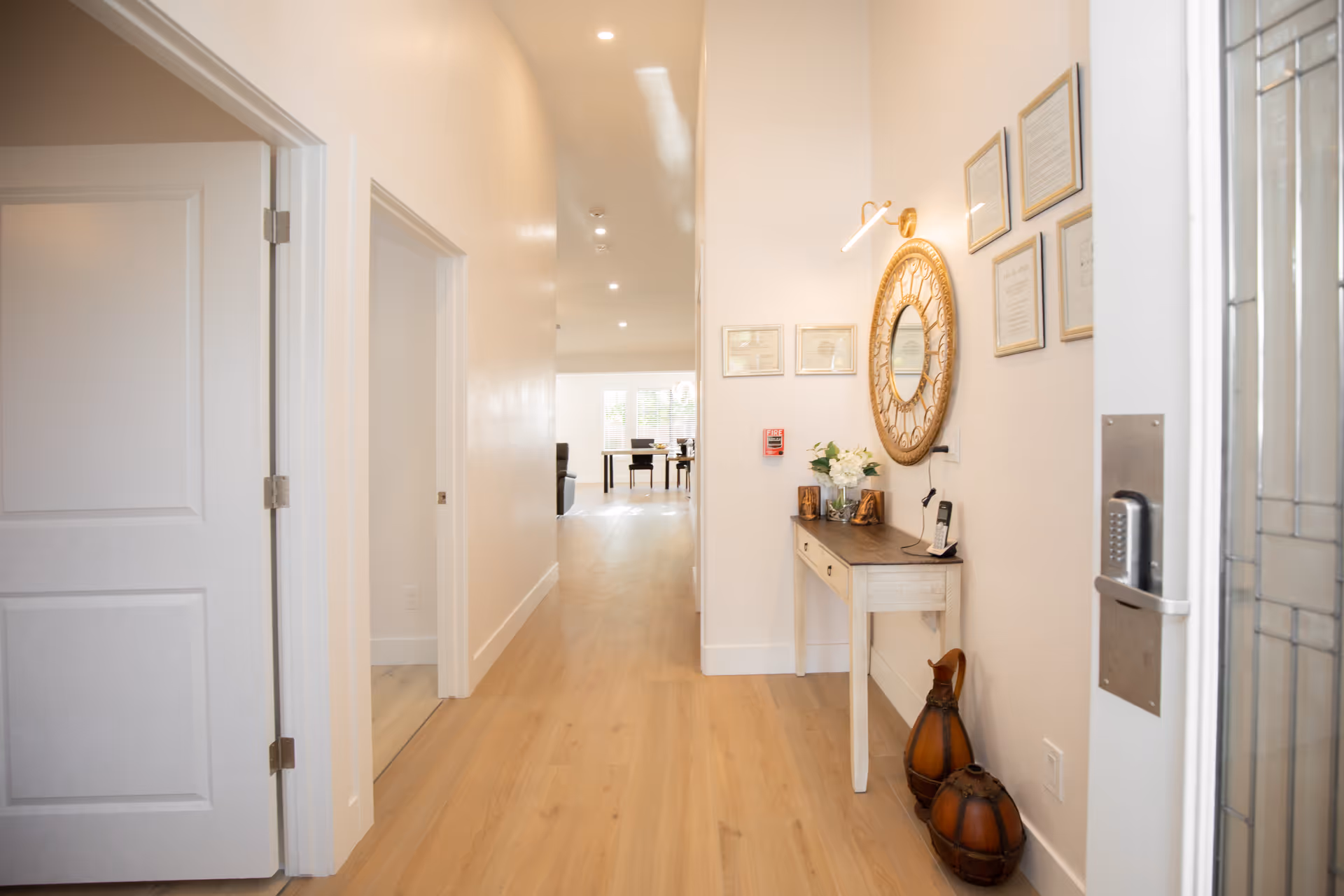 A bright hallway with light wooden flooring and white walls. On the right side, there is a small white console table with decorative items including flowers and two brown vases on the floor. Above the table is a round ornate mirror and several framed pictures on the wall. The hallway leads to a well-lit room with tables and chairs visible in the distance. A door with a keypad lock is partially visible on the right edge of the image.