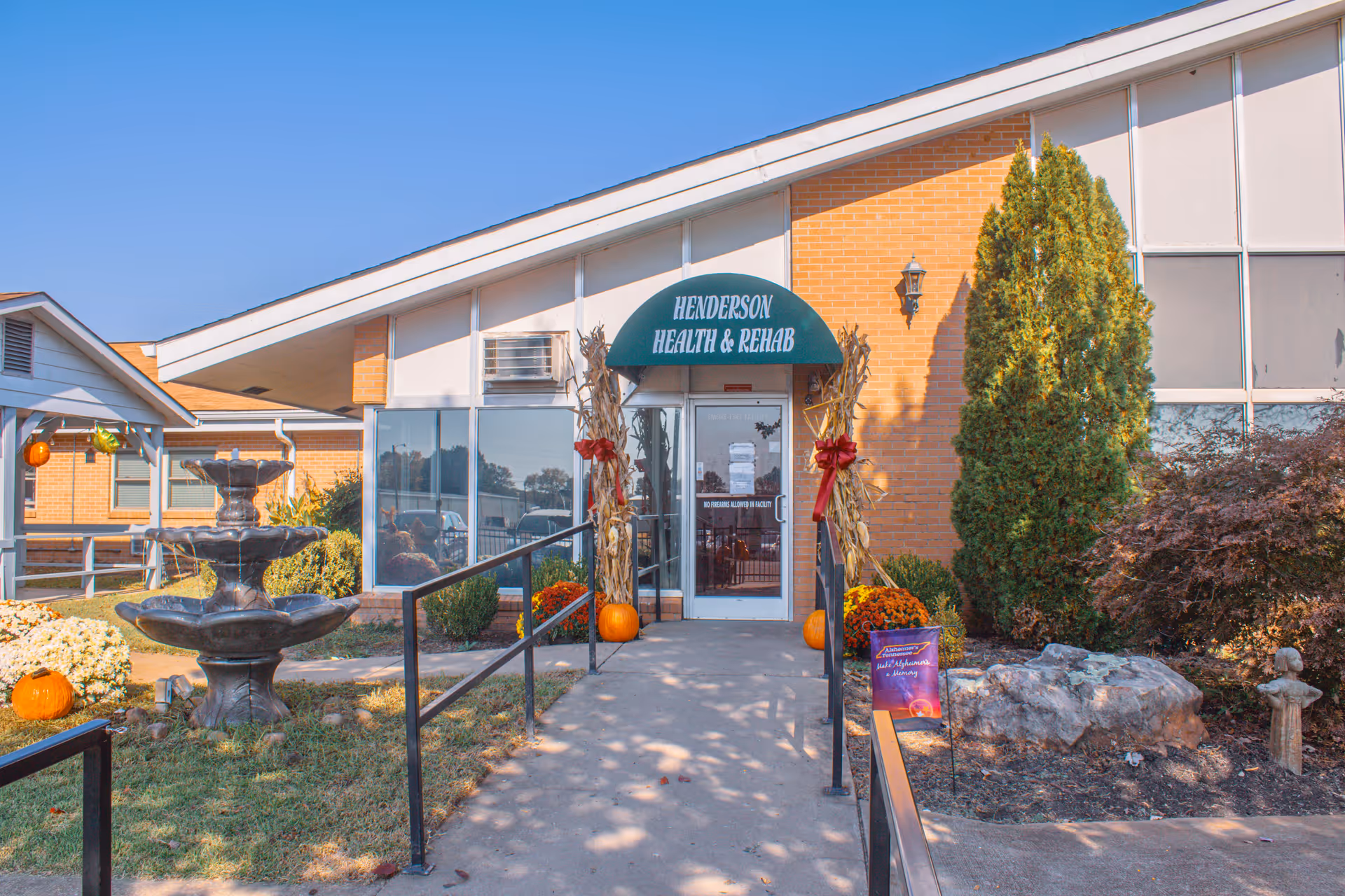 Front entrance of Henderson Health & Rehab with seasonal decorations including pumpkins and corn stalks, a walkway and a fountain in the landscaped foreground.