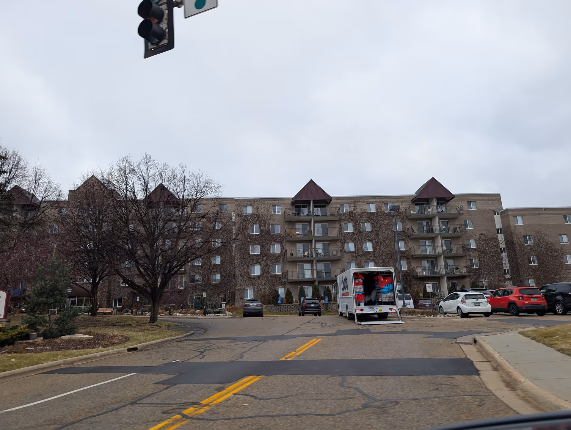 Front exterior of a multi-story senior living building with balconies, bare trees, and parked cars including an open moving truck.