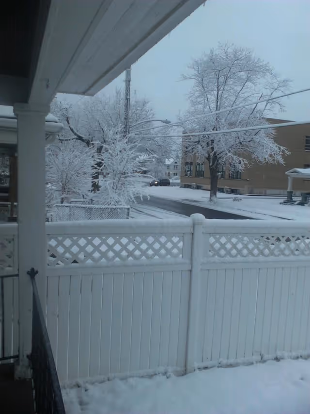 Snow-covered porch view with white fence and bare trees near a building