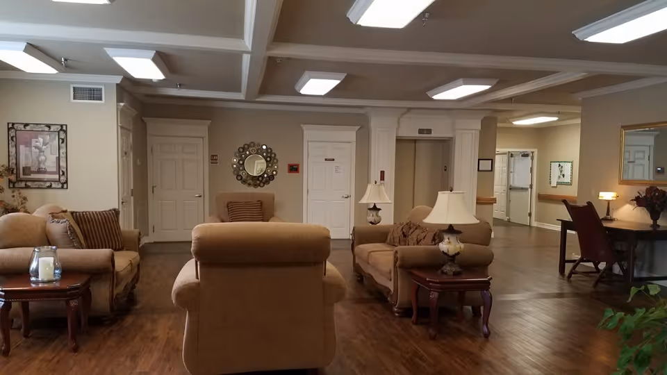 A spacious and well-lit common area in a retirement center featuring comfortable beige armchairs and sofas arranged around wooden side tables with lamps and decorative items. The room has wooden flooring, neutral-colored walls, ceiling lights, and doors leading to other parts of the facility. A round decorative mirror and framed artwork adorn the walls.