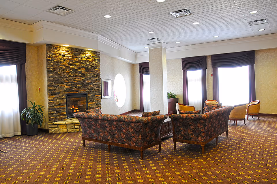 A spacious living room area with patterned carpet, floral upholstered sofas arranged around a wooden coffee table, a stone fireplace with a fire burning, large windows with dark curtains, and potted plants.