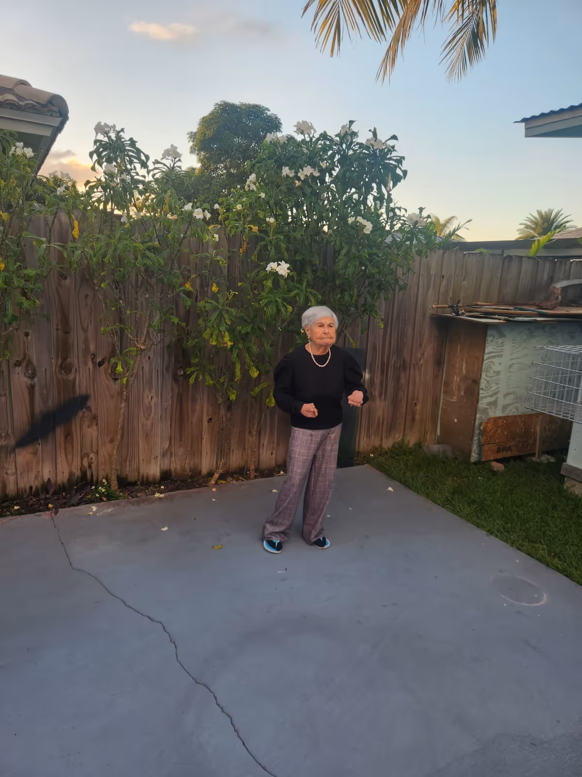 Older woman stands on a concrete outdoor patio beside a wooden fence with flowering shrubs.