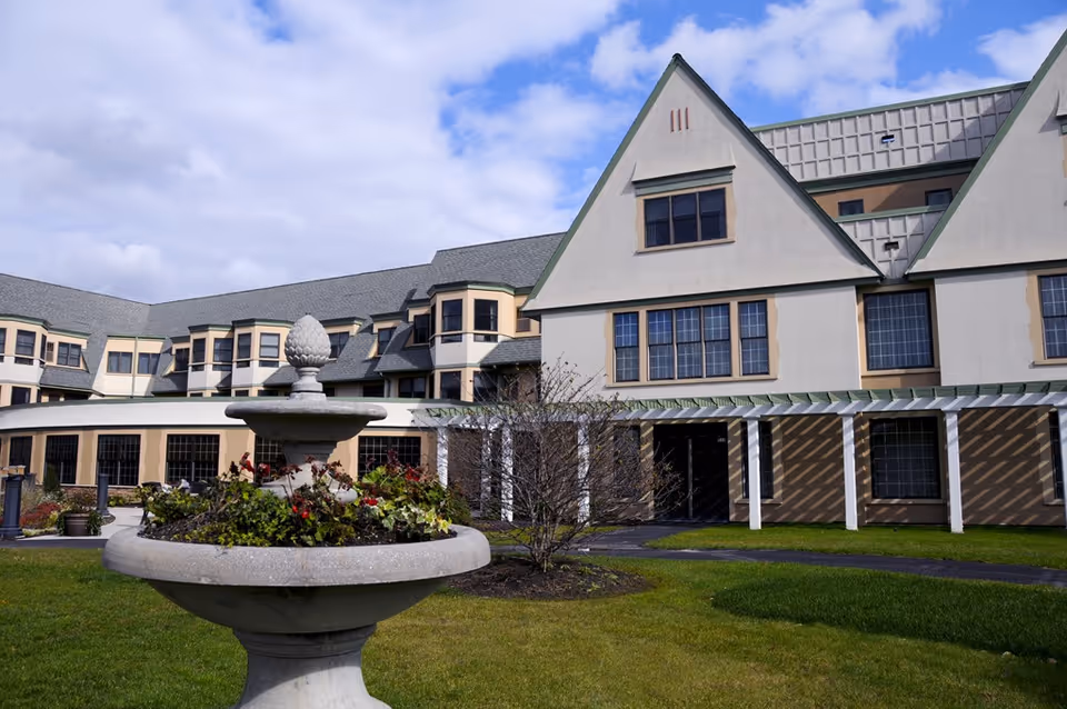 Exterior view of a large multi-story building with beige walls and green trim, featuring multiple windows and a sloped roof. In the foreground, there is a stone planter filled with greenery and flowers on a well-maintained lawn under a partly cloudy sky.