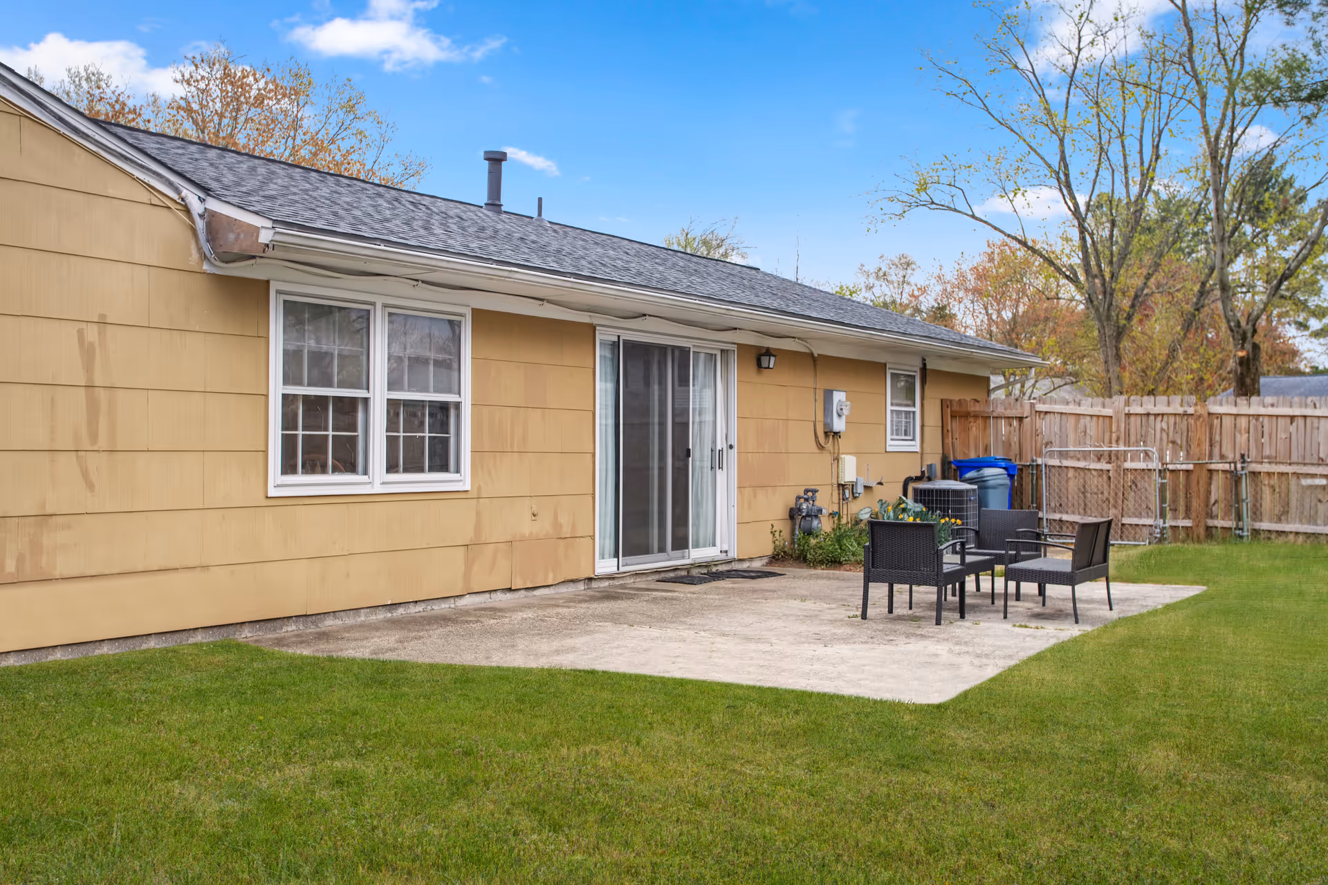 Outdoor patio area of a single-story building with beige siding, featuring a concrete slab with four black chairs and a small table. The area is surrounded by a green lawn and a wooden fence, with trees in the background under a blue sky.