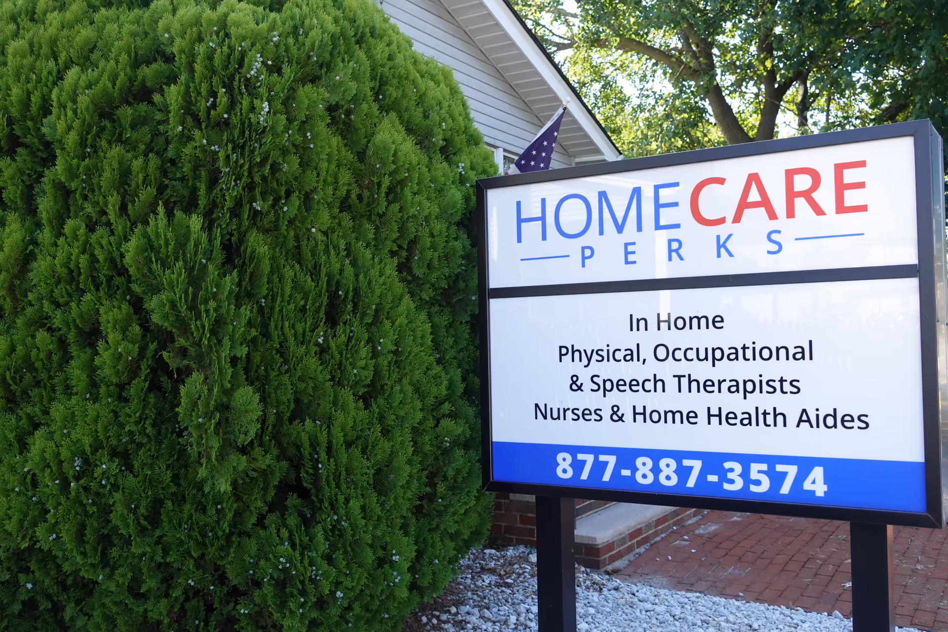 An outdoor sign for HomeCare Perks stands beside tall green shrubs.