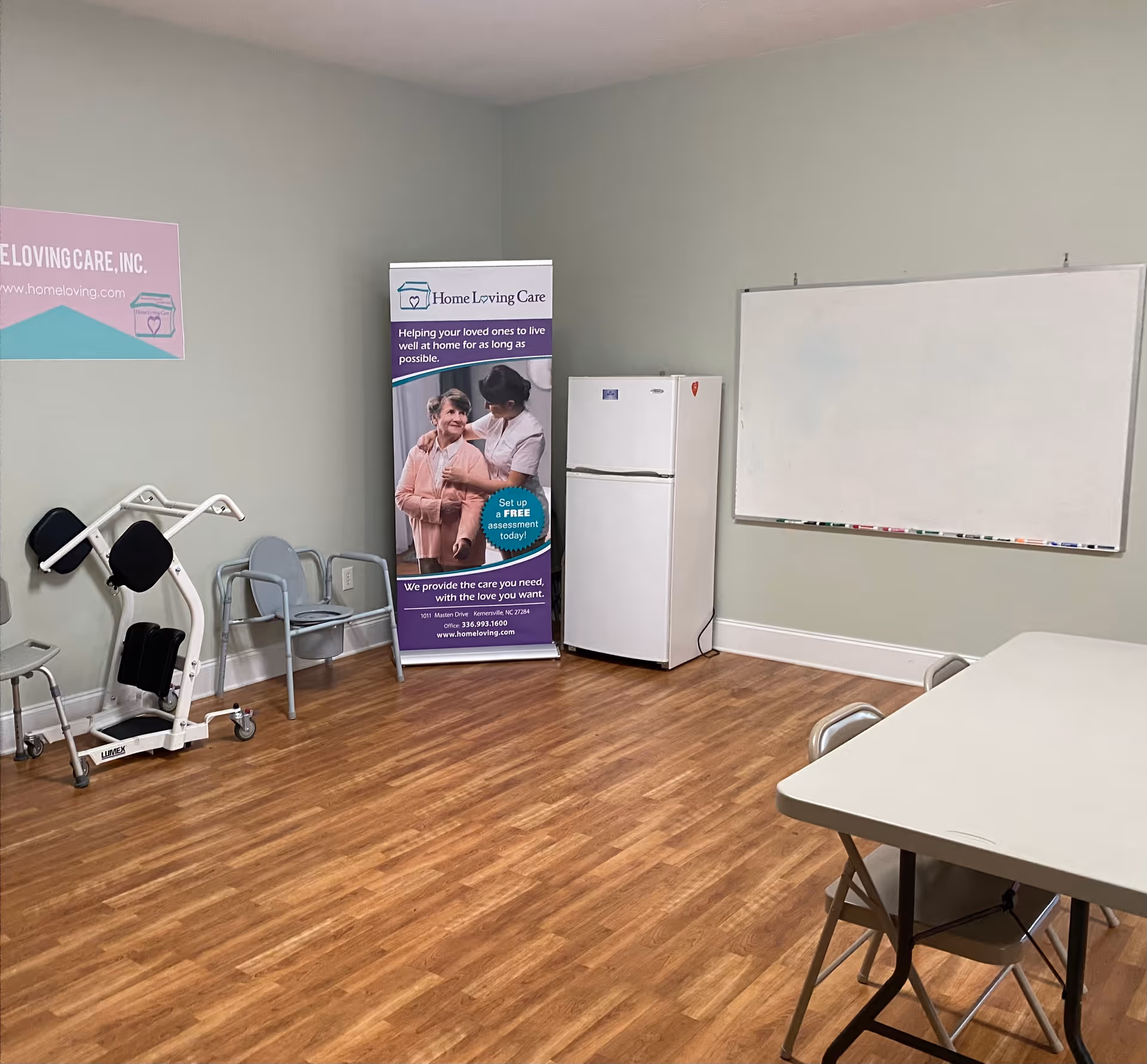 Interior room at Home Loving Care with banner, refrigerator, whiteboard, tables, and chairs.