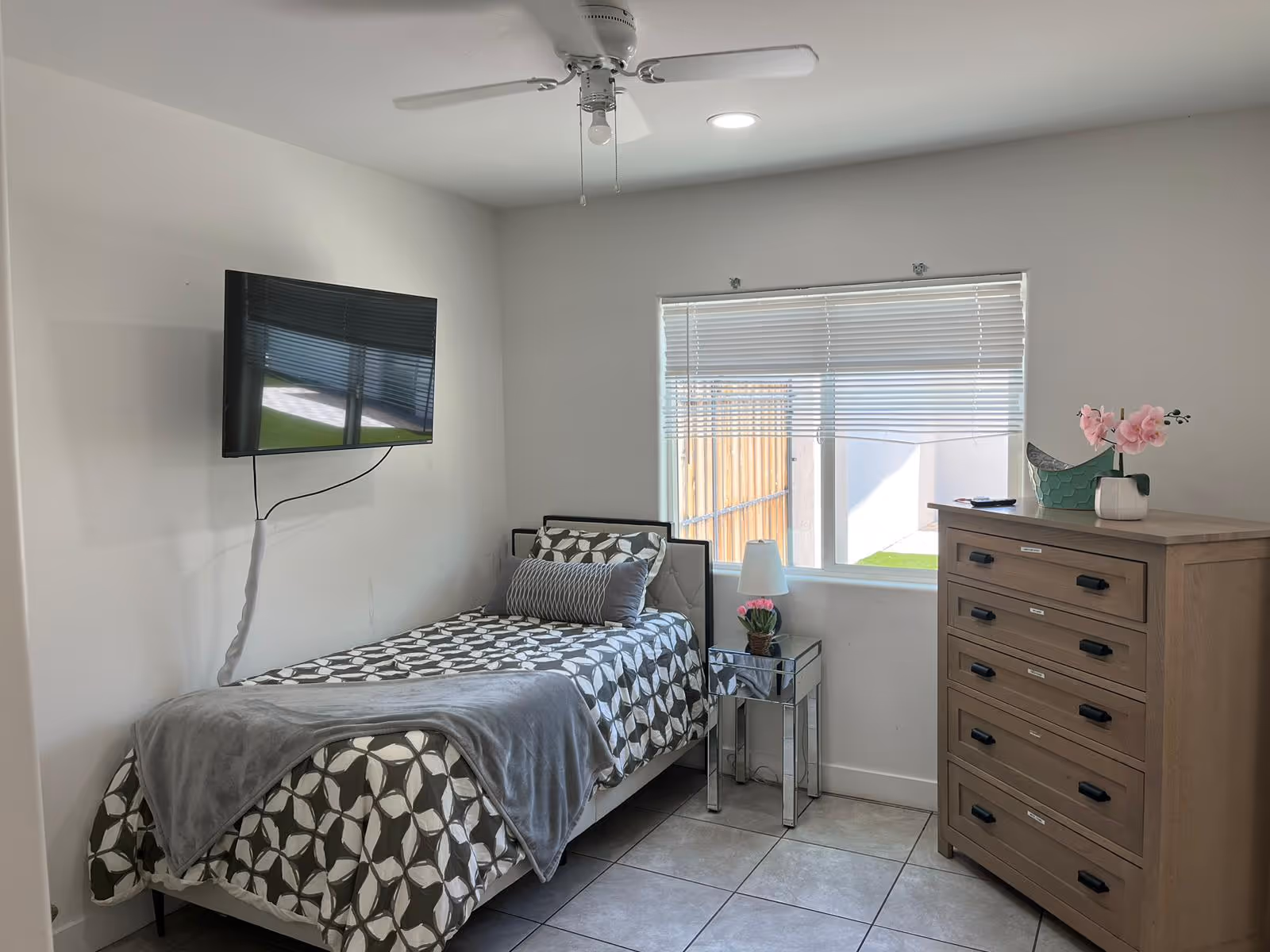 Small bedroom with a single bed, dresser, window blinds, and wall-mounted TV.