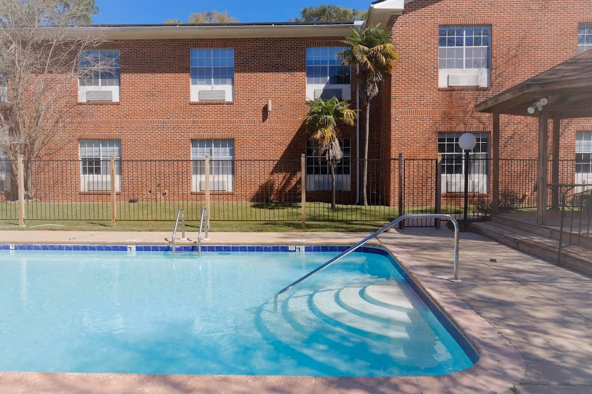Outdoor swimming pool beside a red brick nursing facility, with a fence and palm trees.