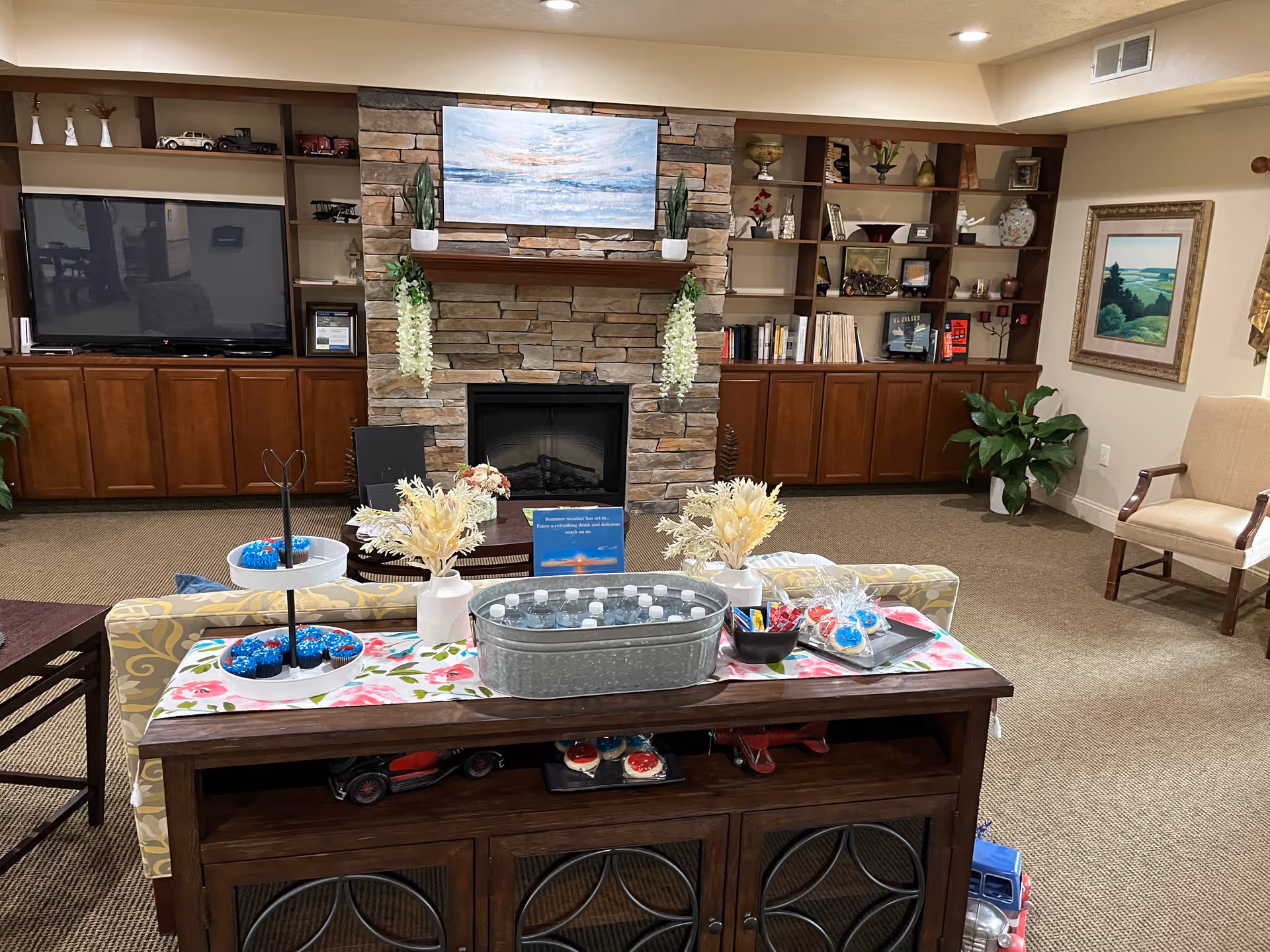 A cozy living room area in an assisted living facility featuring a stone fireplace with a painting above it, flanked by built-in wooden shelves and cabinets filled with decorative items and books. In front of the fireplace is a wooden console table with a floral runner, holding a metal tub filled with bottled water, trays of decorated cookies, and vases with dried flowers. There is a large flat-screen TV on the left side, a beige armchair on the right, and carpeted flooring throughout.