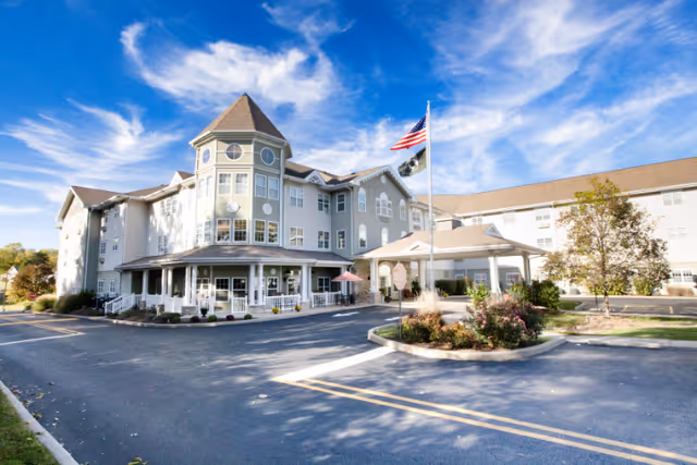 Exterior view of a large senior living facility building with multiple stories, a turret, and a covered entrance. There is an American flag on a flagpole near the entrance, surrounded by landscaped bushes and trees. The sky is blue with scattered clouds.